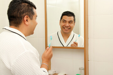 Dark-skinned 40-year-old Latin man brushes his teeth in front of the bathroom mirror in a bathrobe...