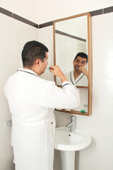 Dark-skinned 40-year-old Latin man brushes his teeth in front of the bathroom mirror in a bathrobe...