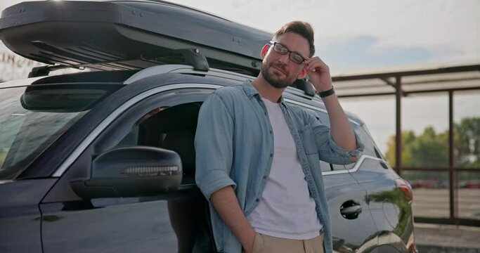 Young Caucasian man standing near the car with roof racks and cargo box on a parking lot and looking at camera