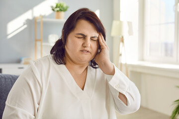 Overweight woman suffering from sudden attack of headache or dizziness. Portrait of exhausted plus size woman sitting on sofa touching her temple feeling fatigue, nervous tension