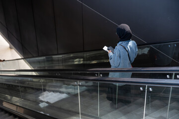 Young casual female using escalator in airport holding her passport and using her mobile phone...