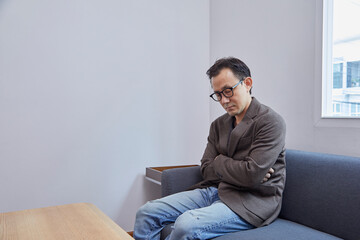 Asian businessman wearing eyeglasses in dark suit, taking a nap on grey sofa in modern living room, tired and exhausted from busy day at workplace.