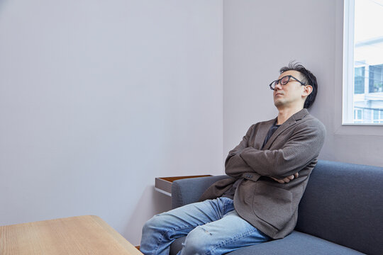 Asian businessman wearing eyeglasses in dark suit, taking a nap on grey sofa in modern living room, tired and exhausted from busy day at workplace. - Powered by Adobe
