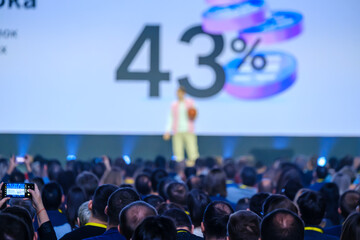 Business crowd listening to male speaker on stage with number and percentage sign being displayed...