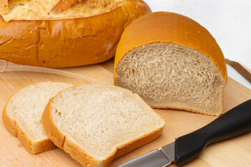 bakery products. bread cut into slices with a black knife lies on a light wooden board, on a white wall background