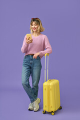 Happy girl tourist holding travel suitcase using mobile phone standing isolated on purple...