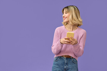 Happy pretty gen z blonde young woman model holding smartphone looking at copy space aside, smiling girl using mobile apps on cell phone standing isolated on purple background with cellphone. - obrazy, fototapety, plakaty
