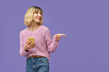 Happy pretty gen z blonde young woman model holding smartphone pointing at copy space aside, smiling girl using mobile apps on cell phone standing isolated on purple background with cellphone. - obrazy, fototapety, plakaty