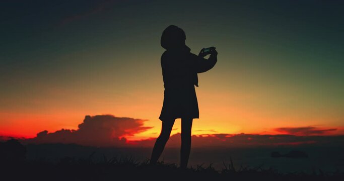 Girl silhouette captures sunset sky nature beauty. Female make photo on her phone standing over sea, enjoy bright colorful sky. Outdoor lifestyle travel, summer holiday vacation. Back view slow motion