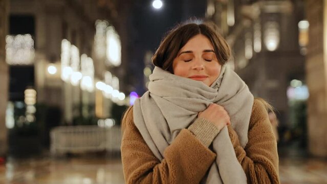 Woman covers her body with a scarf and close her eyes. Woman wearing warm clothing. Steadicam shot on the night street of Milan, Italy
