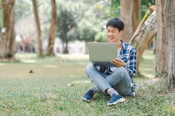 Young Asian student is sitting on the grass, focused on his laptop screen, probably working on his homework or studying.