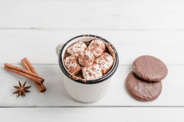 Homemade spicy hot chocolate drink with marshmallows in enamel cup on white wooden table with cinnamon stick, cookies and star anise