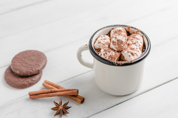 Homemade spicy hot chocolate drink with marshmallows in enamel cup on white wooden table with cinnamon stick, cookies and star anise