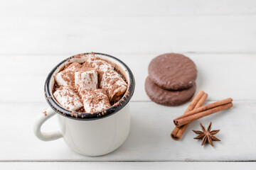 Homemade spicy hot chocolate drink with marshmallows in enamel cup on white wooden table with cinnamon stick, cookies and star anise