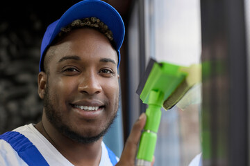 Smiling cute curly guy in a blue baseball cap wipes windows from dust with a scraper with a rubber band for windows. Close up of the happy face of a black blue-uniformed worker cleaning the premises.  - obrazy, fototapety, plakaty