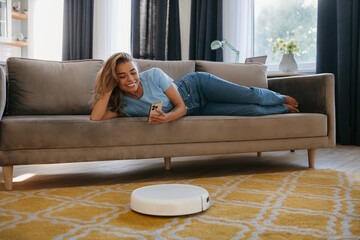 Happy young woman relaxing on the couch and using mobile phone to control smart vacuum cleaner robot