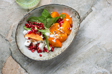 Bowl of fresh persimmon, goat cheese, spinach and pomegranate salad, above view on a grey granite...