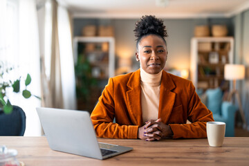 Successful stylish female freelancer looking at camera sitting at home office desk with laptop computer and coffee cup.