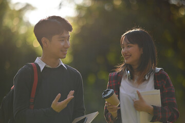 Backlit photograph of two Asian students talking while walking in a park - obrazy, fototapety, plakaty