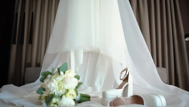 Close-up of the bride's white wedding dress. Huge spacious room. Bridal bouquet and shoes on the bed for the bride. The bride's formal morning at the hotel.