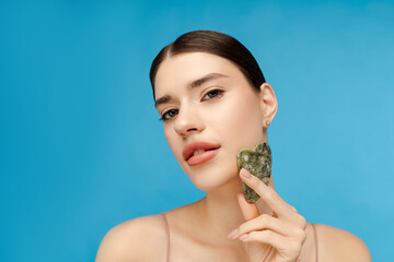 Happy woman doing skin care routine, massaging her cheekbone with jade gua sha scrapper. Isolated on blue background. Anti-Aging skincare routine