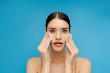 Pretty young woman using cotton pads and cleansing face skin. Isolated on blue background....