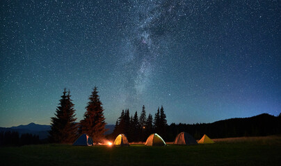 Night camping in mountains under starry sky. Tourist tents in campsite near burning campfire under beautiful sky full of stars with Milky way above forest. Concept of tourism and traveling.