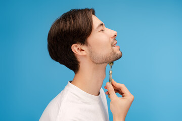 Side view of smiling man massaging his chin with jade roller in studio. Isolated on blue...