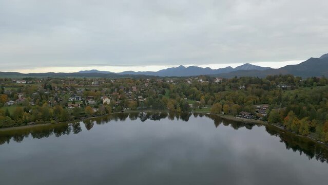 Descending aerial footage above the peaceful and placid waters of Staffelsee, reflecting trees and scenery against the backdrop of the German Alps and the town of Murnau