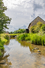 Fototapeta na wymiar Low angle perspective of Geul river, old Eper watermill or Wingbergermolen against gray sky in background, wild vegetation and lusch trees, cloudy day in Terpoorten, Epen, South Limburg, Netherlands