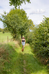 Naklejka na ściany i meble Adult female hiker in summer clothes walking on path with dog between farm agricultural plots, plants and leafy trees in background, shorts, sunglasses, sunny day in Epen, South Limburg, Netherlands