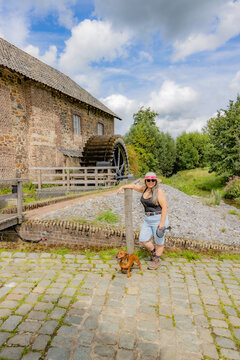 Smiling mature woman with her brown dachshund standing in front of old Eper or Wingbergermolen water mill in background, sunny day blue sky with clouds in Terpoorten, Epen, South Limburg, Netherlands