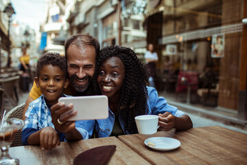 Happy multiracial couple taking selfie with little son sitting in outdoor cafe