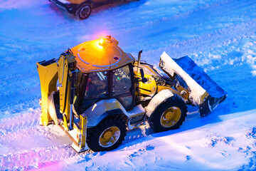 A tractor loader in selective focus is going to remove snow from the road at night. A snowplow cleans the driveway, yards of a house in the city after a heavy snowfall and blizzard.
