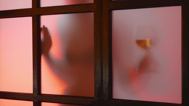 Enigmatic picture of unrecognized nude model with a glass of wine behind matt glass at sunset. Seductive girl, young flexible lady with perfect body, moving silhouette. Slow mo high quality 4k footage