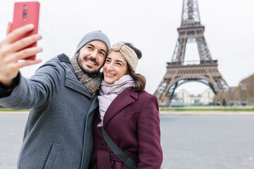European young people enjoying winter vacation in France. Spanish adult couple taking selfie portrait together in front of Eiffel Tower in Paris. Tourism and holidays concept.