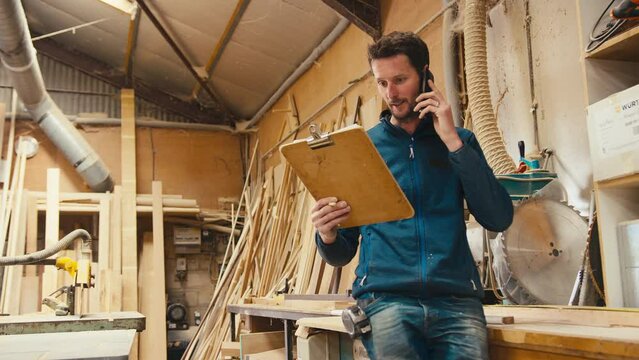 Male carpenter working in woodwork workshop making call on mobile phone whilst looking at plan on clipboard - shot in slow motion