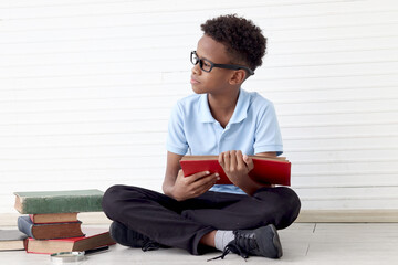 Serious concentrate African boy wearing glasses while sitting on floor with book stack in white wall room. Cute child with pile of books, kid education, learning, reading and exploring discovery.