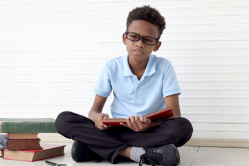 Concentrate African boy with glasses using magnifying glass to read book while sitting on floor in white wall room. Cute child with pile of books, kid education, learning, exploring discovery.