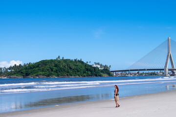 mujer caminando por la playa con un puente colgante de fondo y una montaña 