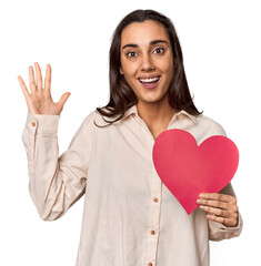 Caucasian young woman with paper heart for Valentine's receiving a pleasant surprise, excited and raising hands.