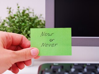 A person hand sticking a green paper note with the reminder Now or Never on it on to a monitor at...
