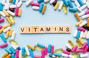 A variety of colorful pills and capsules scattered on a bright blue background. The word VITAMINS is spelled out using wooden tiles.