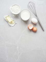 Fototapeta premium Ingredients for homemade pastry: flour, milk, eggs and butter on gray stone table, hand metal whisk. Top view with copy space.
