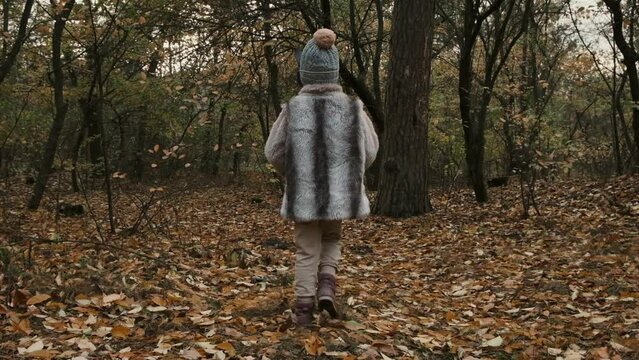 Happy little girl walking in a slow motion through the autumn forest in the mask of wolf and wearing fur. View from behind.