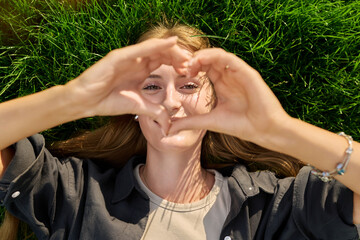 Young happy loving woman showing heart sign, looking at camera lying on grass