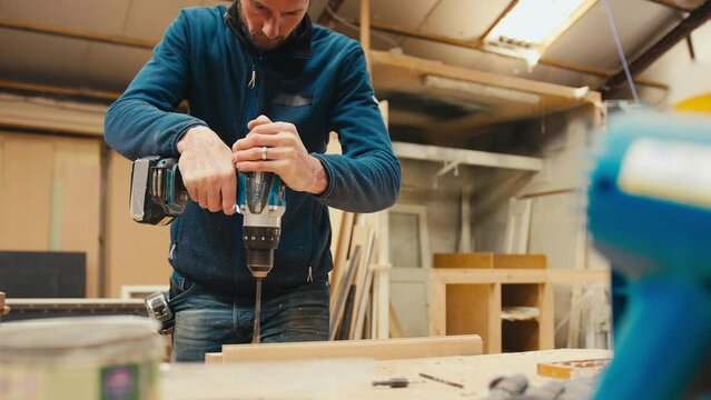 Male carpenter at workbench drilling hole in piece of wood with cordless electric drill - shot in slow motion