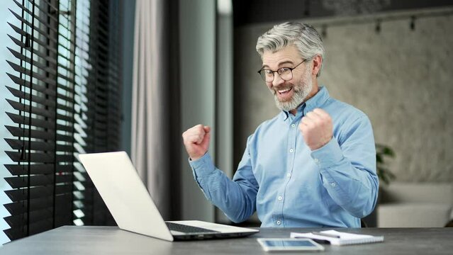 Excited happy mature gray haired bearded businessman received great news on laptop sitting at workplace in business office. Joyful teacher reads a pleasant message, celebrates success surprise yes wow