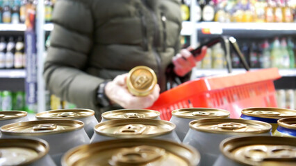 Close-up of many golden-white cans of beer in alcohol department and a man with a shopping basket...