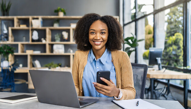 Portrait Of A Beautiful Confident Businesswoman Using A Laptop Computer Holding A Mobile Phone Sitting In A Modern Office. Smiling African American Freelancer Working Online From Home